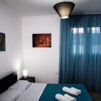 Aegean View Villa 16 Aegean View Villa – Υπνοδωμάτιο με διπλό κρεβάτι, γαλάζιες κουρτίνες και πίνακες τοίχου